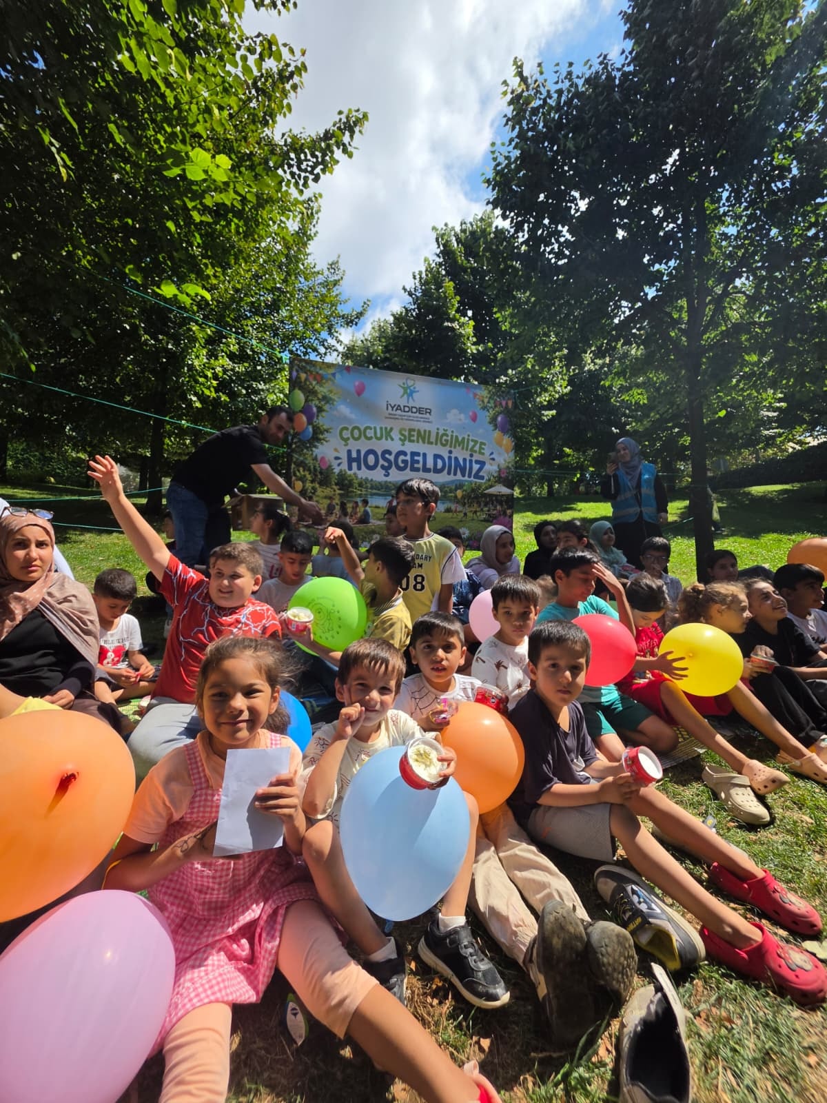 Balonlarıyla çimende oturan çocuklar; arkada çocuk şenliği pankartı