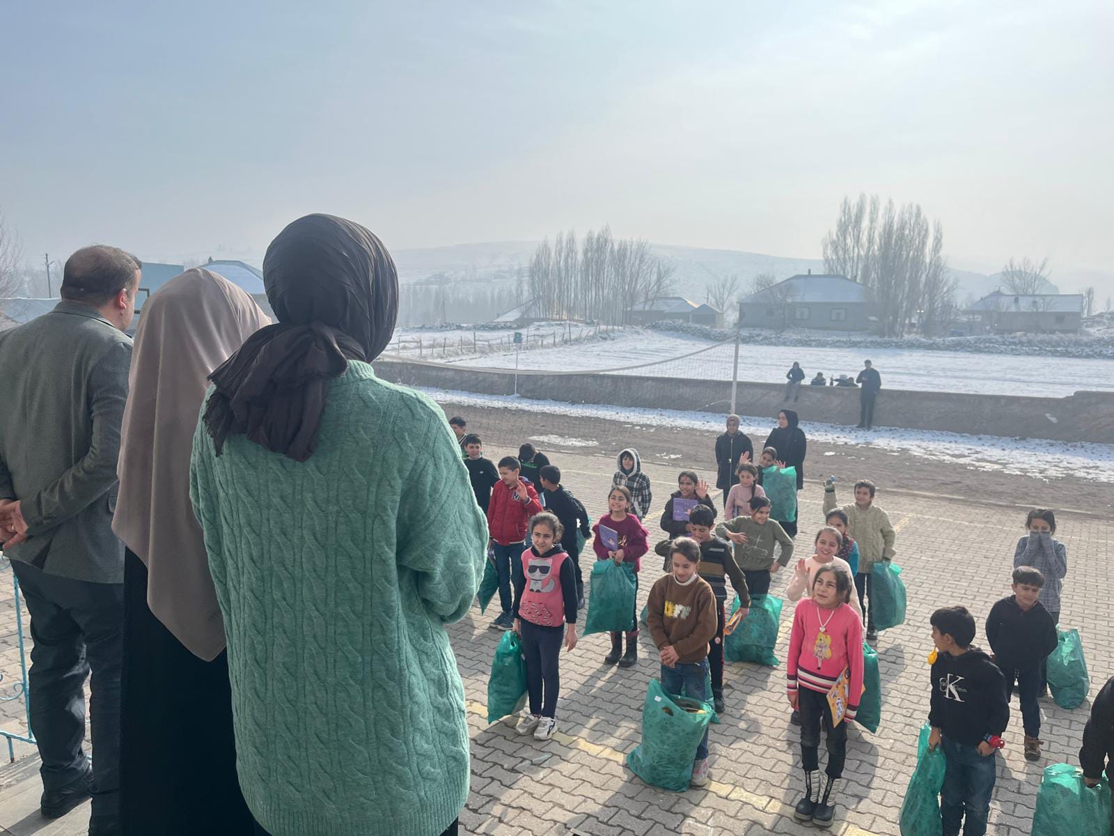 Karlı okul bahçesinde paketlerini alan öğrencilerle konuşan gönüllüler